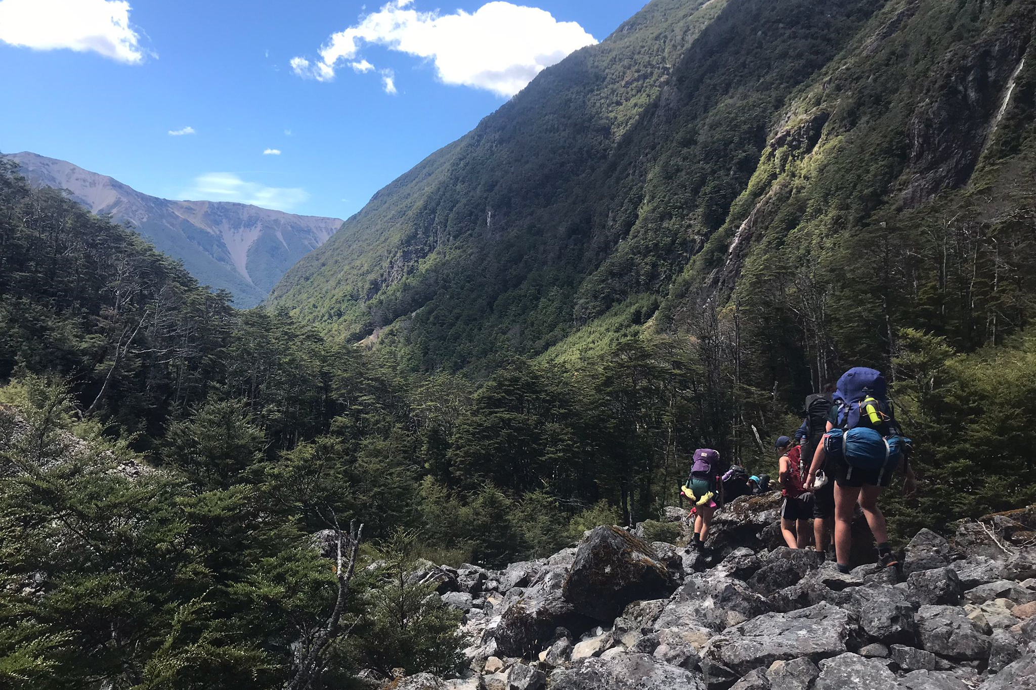 Participants with heavy packs walk over boulders in a valley floor