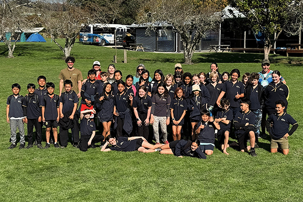a group of 40 students huddle for a photo on a lawn with trees in the background